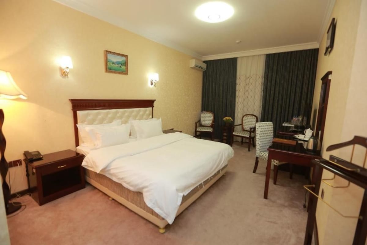 A hotel room with a bed, bedside table, lamp, chair, desk, and a painting on the wall.