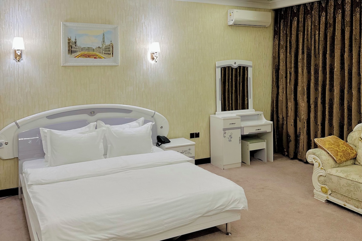 A hotel room with a large bed, bedside tables, a desk, a chair, an air conditioning unit, and a painting on the wall.