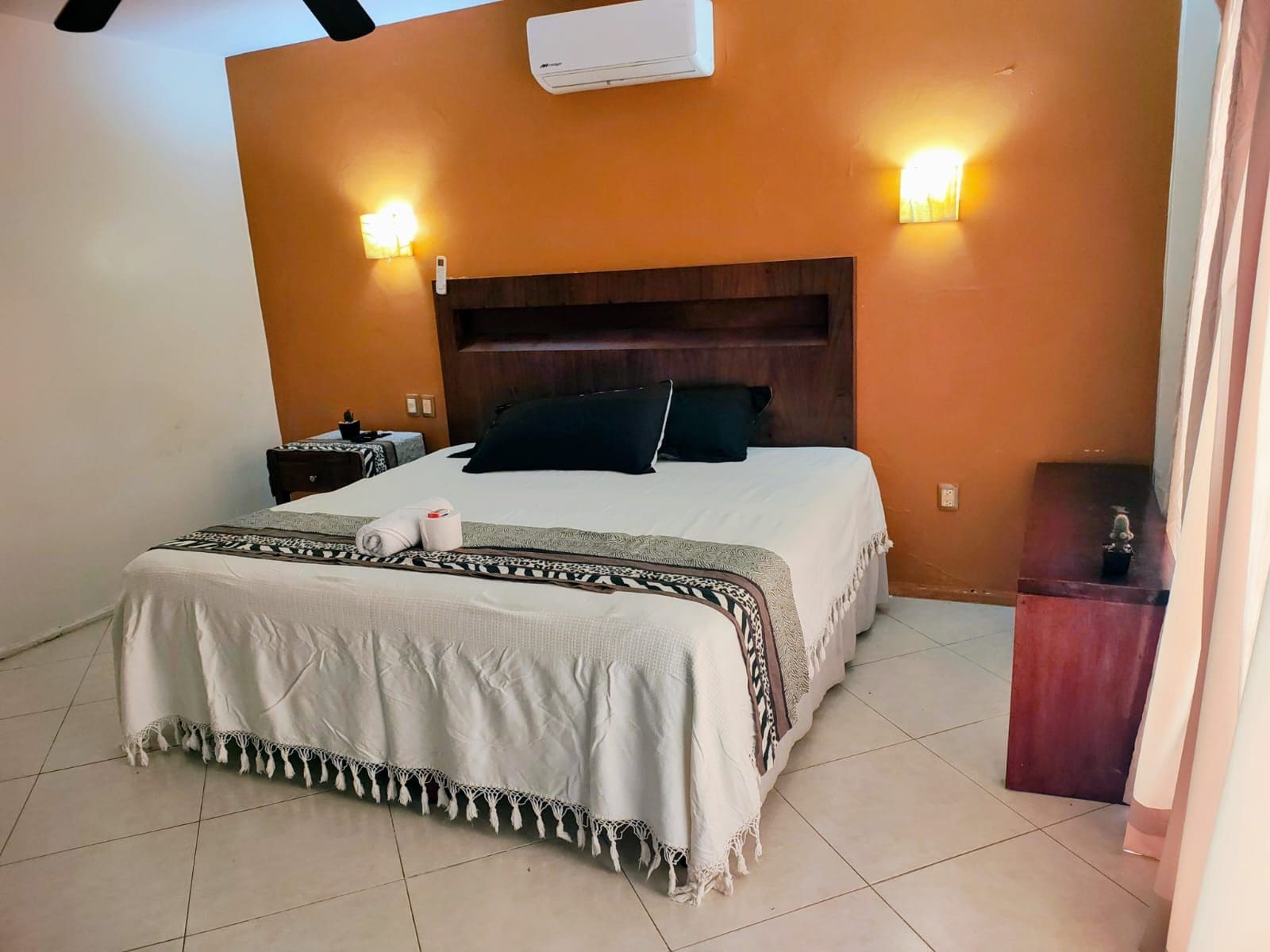 A hotel room with a bed, bedside tables, an air conditioning unit, and wall-mounted lights.