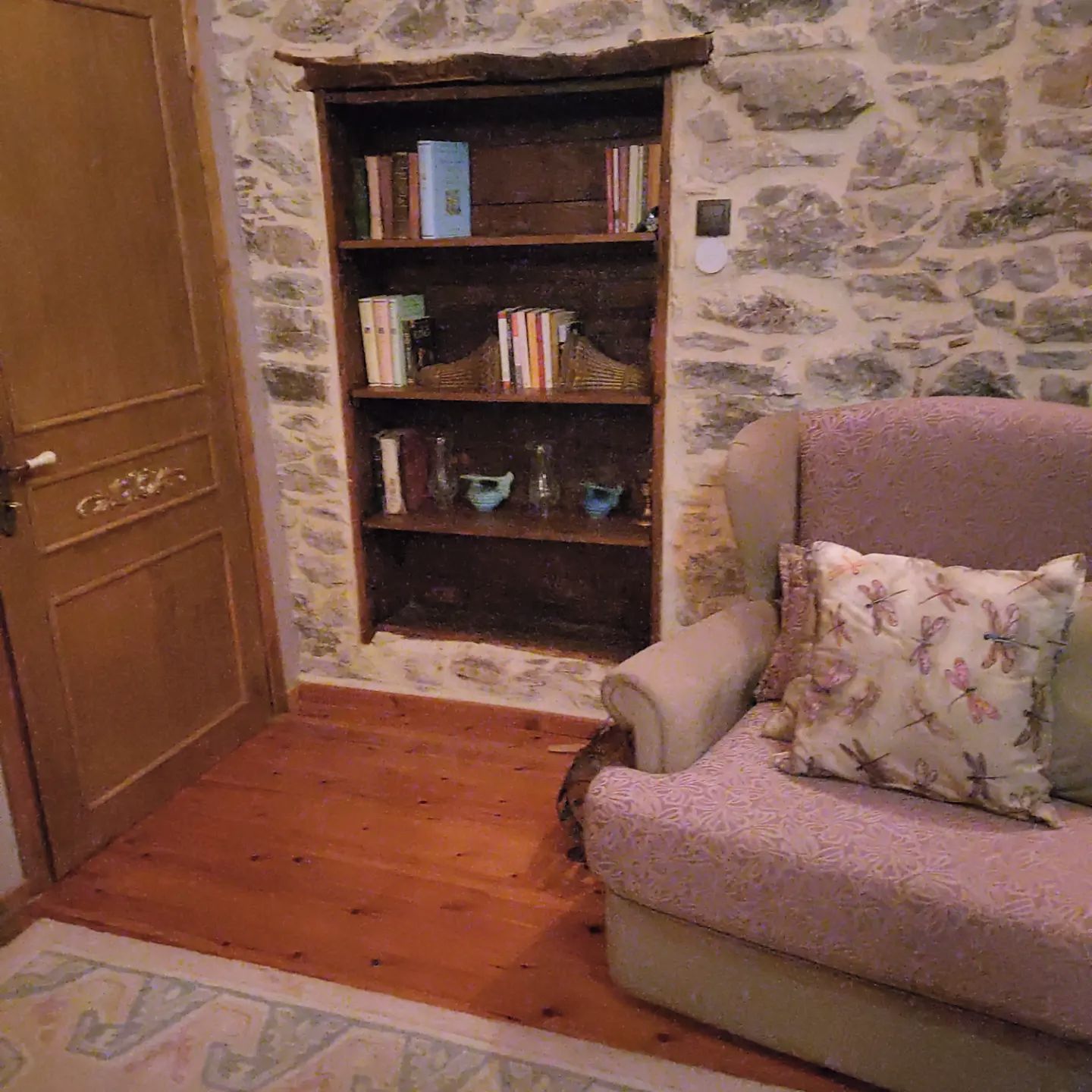 A room with a wooden door, a bookshelf, a patterned armchair, and a stone wall.