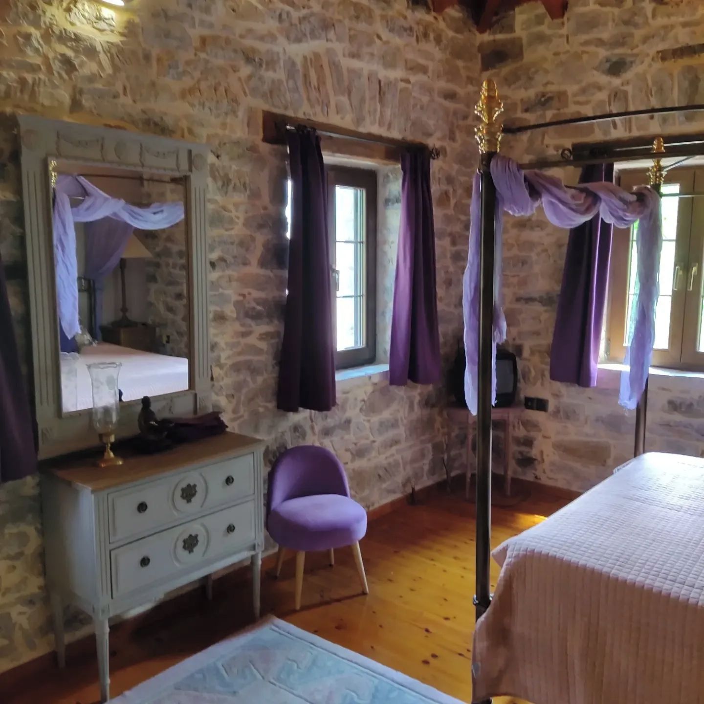 Una habitación con paredes de piedra, una cama, un aparador, una silla y un ventanal con cortinas.
