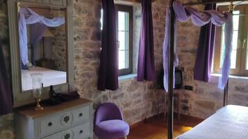 Una habitación con paredes de piedra, una cama, un aparador, una silla y un ventanal con cortinas.