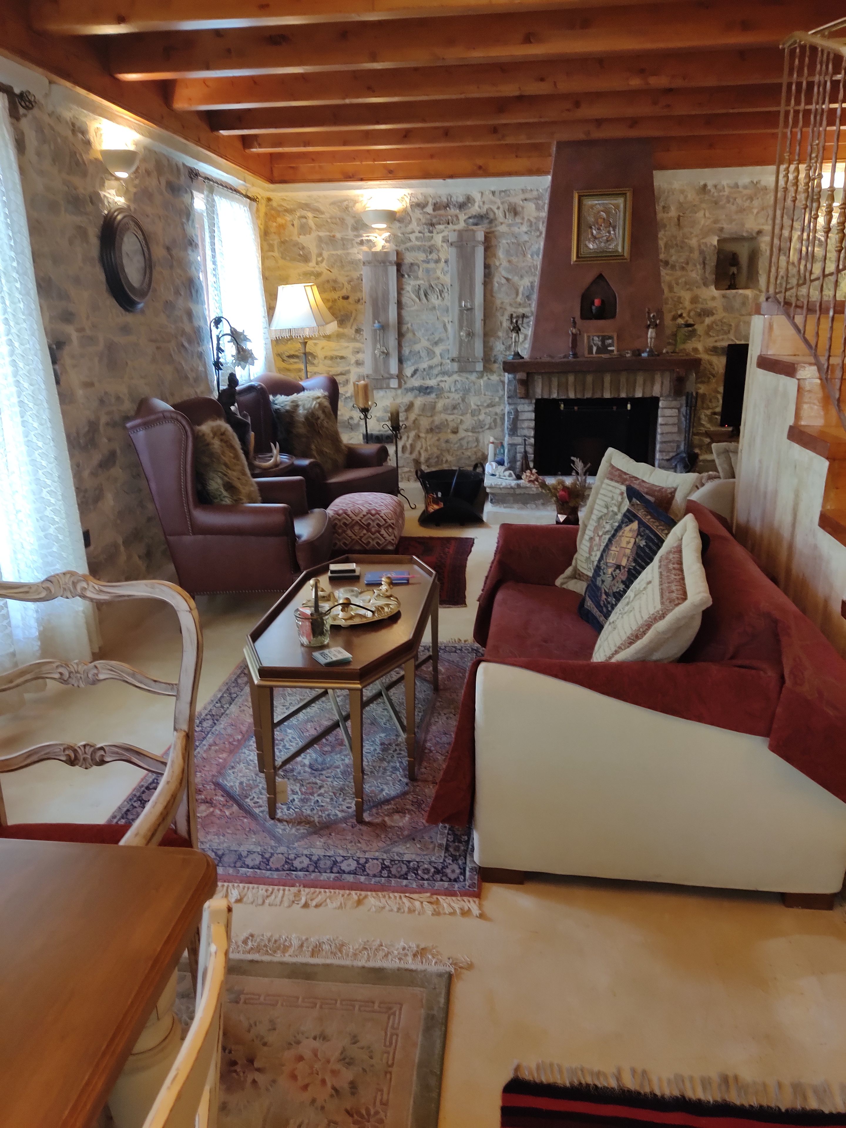 A cozy living room with a fireplace, stone walls, wooden beams, and a mix of furniture including a sofa, armchair, and a coffee table.