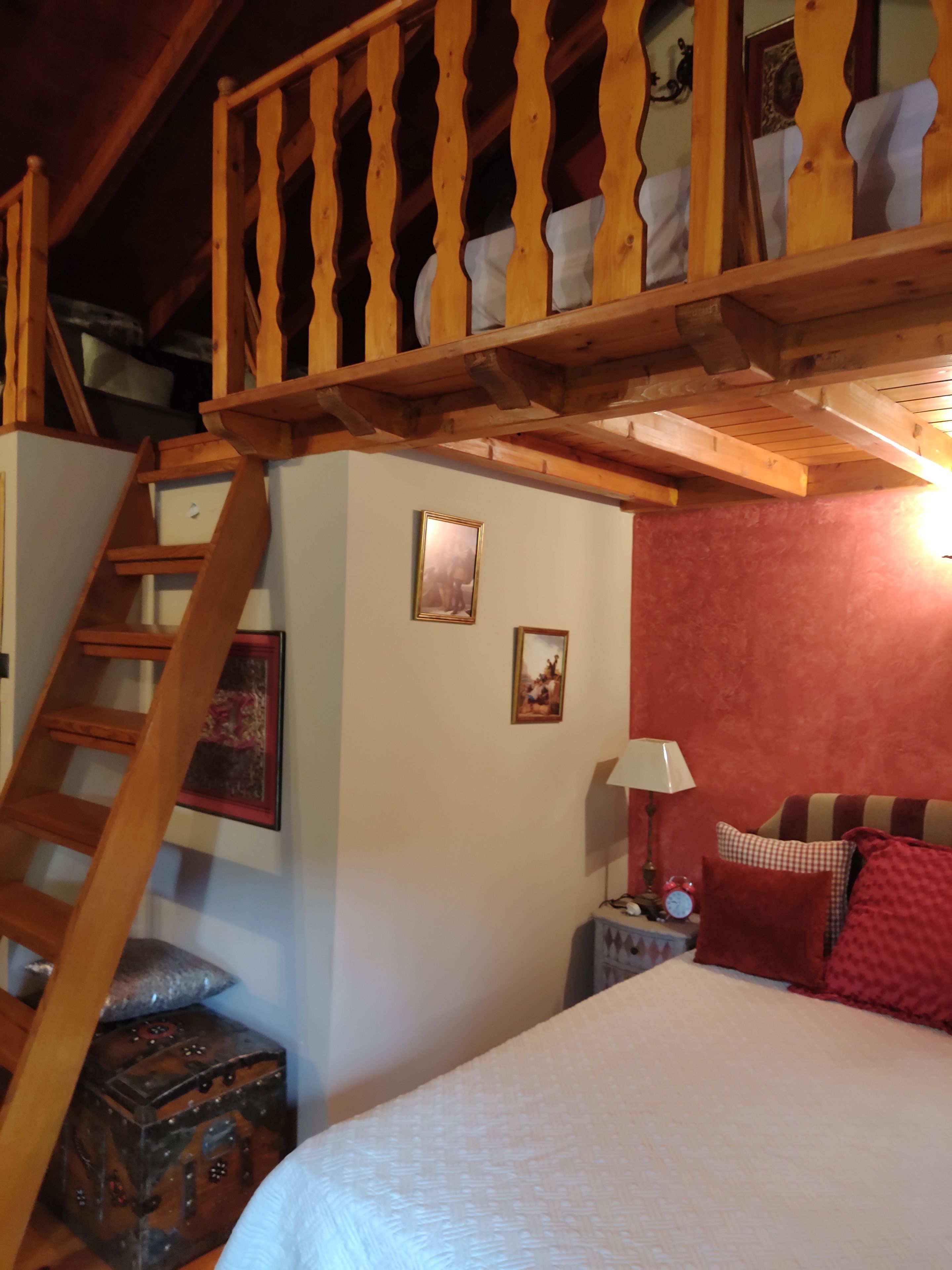 A bedroom with a wooden staircase, a bed, a nightstand with a lamp, and two framed pictures on the wall.