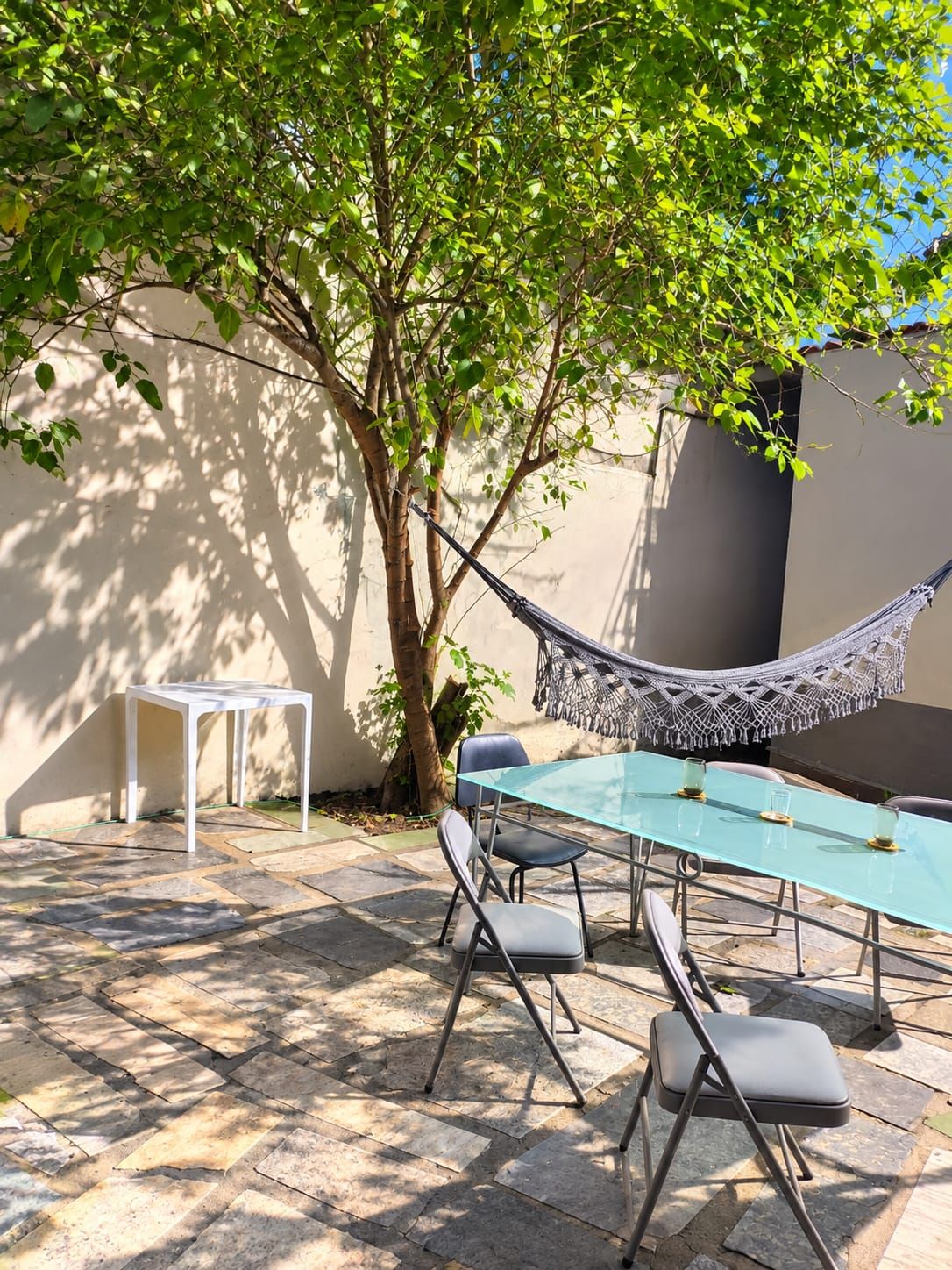 Un patio exterior con una mesa de vidrio, cuatro sillas, una hamaca y una pequeña mesa blanca.