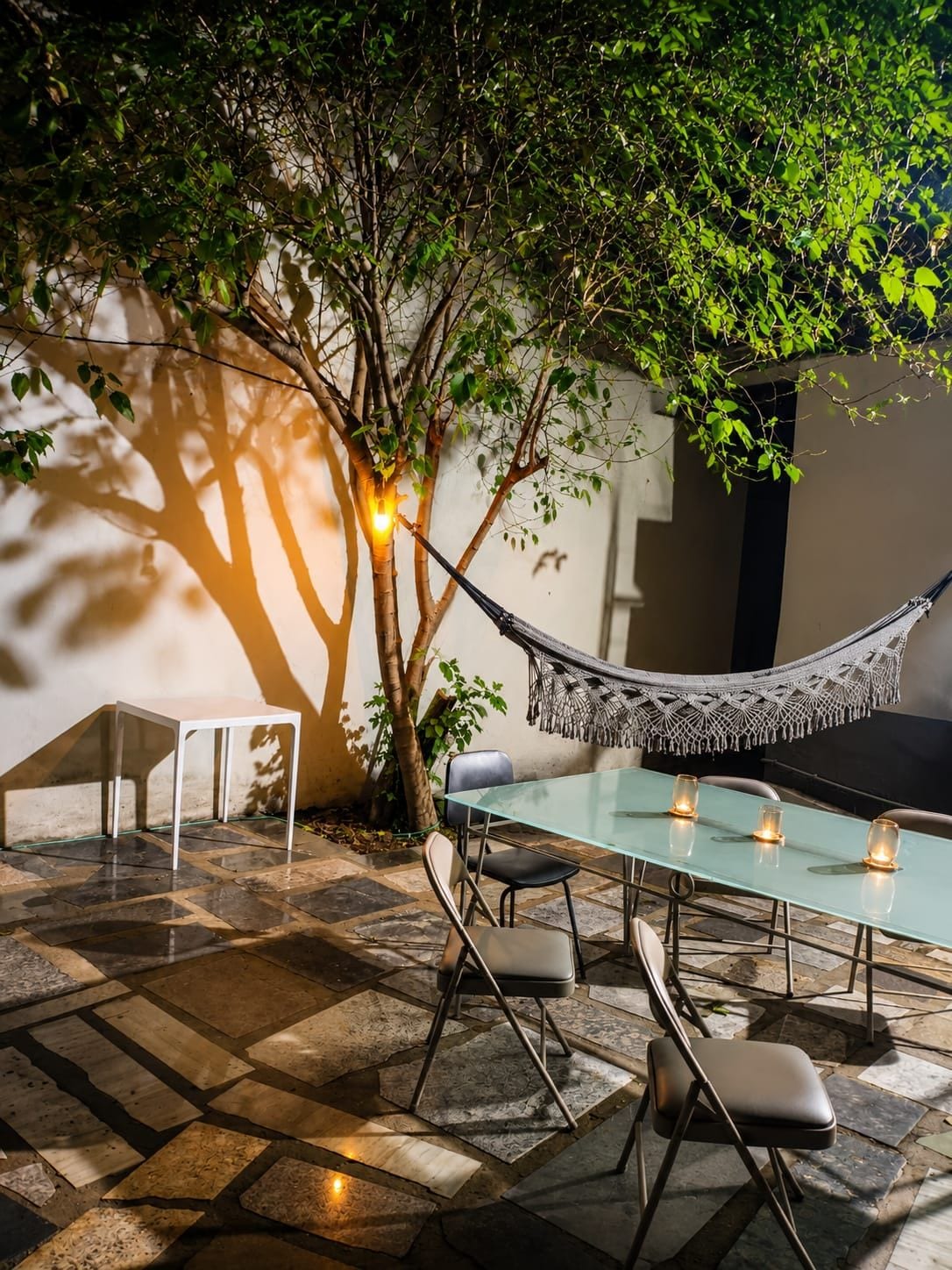 Un espace de restauration en plein air avec une table en verre, des chaises, un hamac et un luminaire fixé au mur.