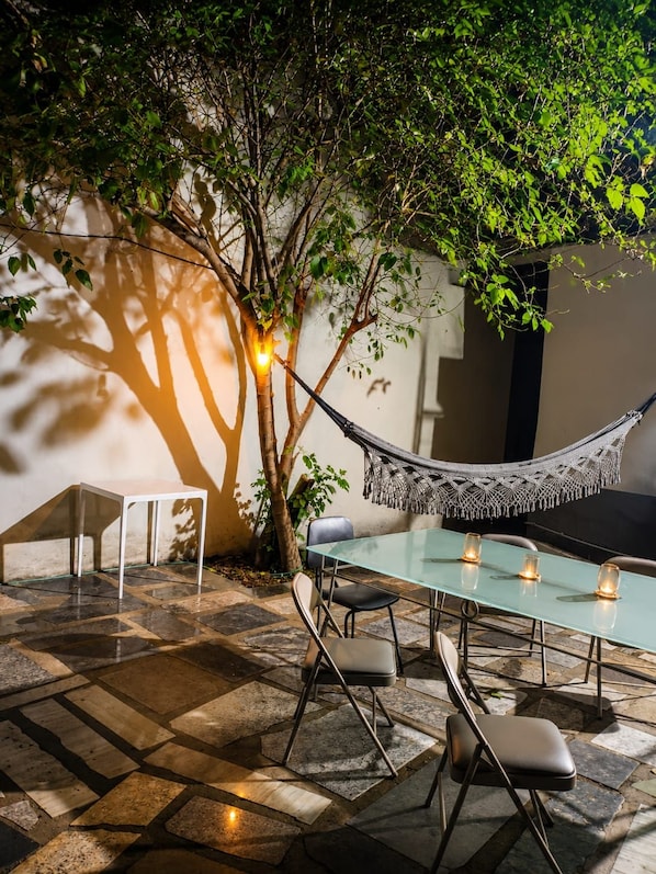 An outdoor dining area with a glass table, chairs, a hammock, and a wall-mounted light fixture.