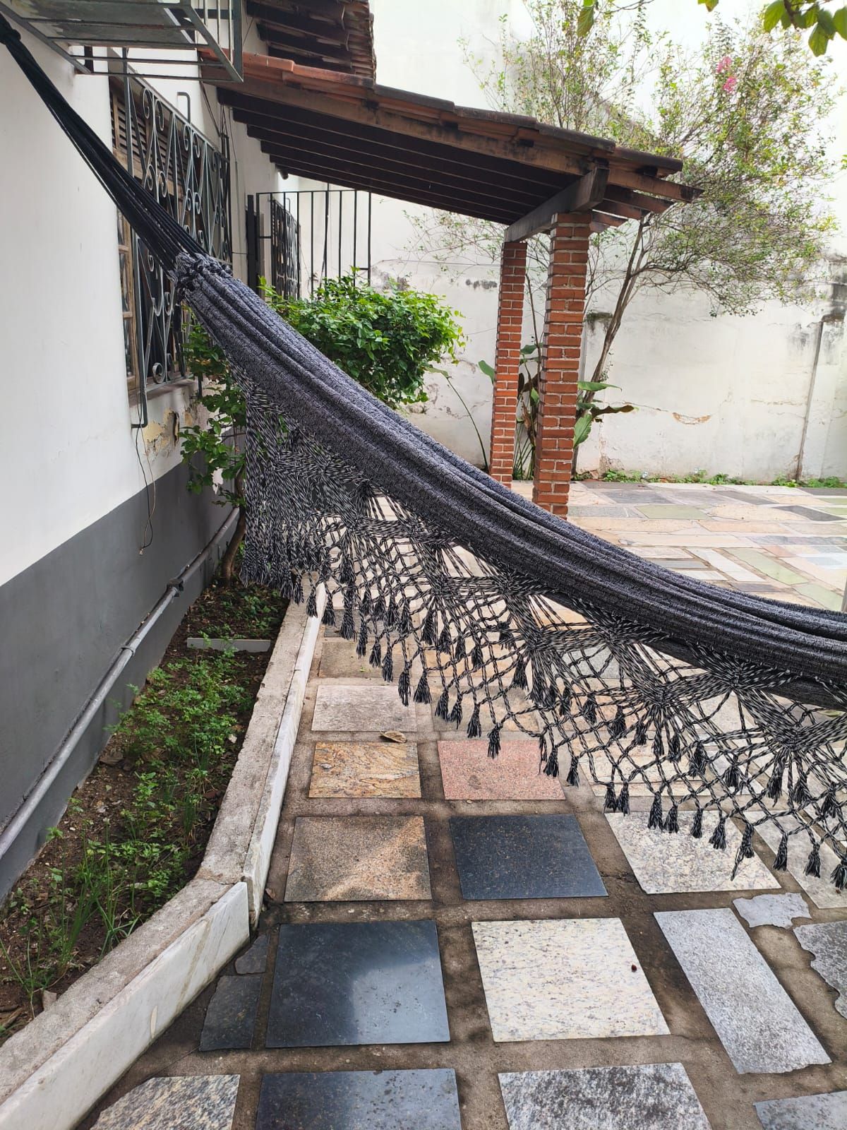 Un hamac est suspendu entre un pilier en briques et un mur blanc, au-dessus d’une terrasse carrelée.