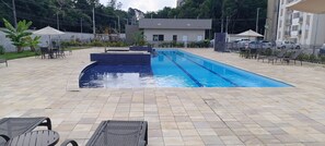 Une piscine dont les bords sont recouverts de carrelage, entourée de chaises longues et de parasols, avec un bâtiment en arrière-plan.