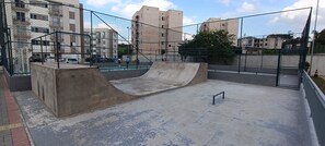 Un parc à roulettes en plein air, doté de rampes en béton et d’un terrain de basketball, le tout entouré d’une clôture en treillis métallique.