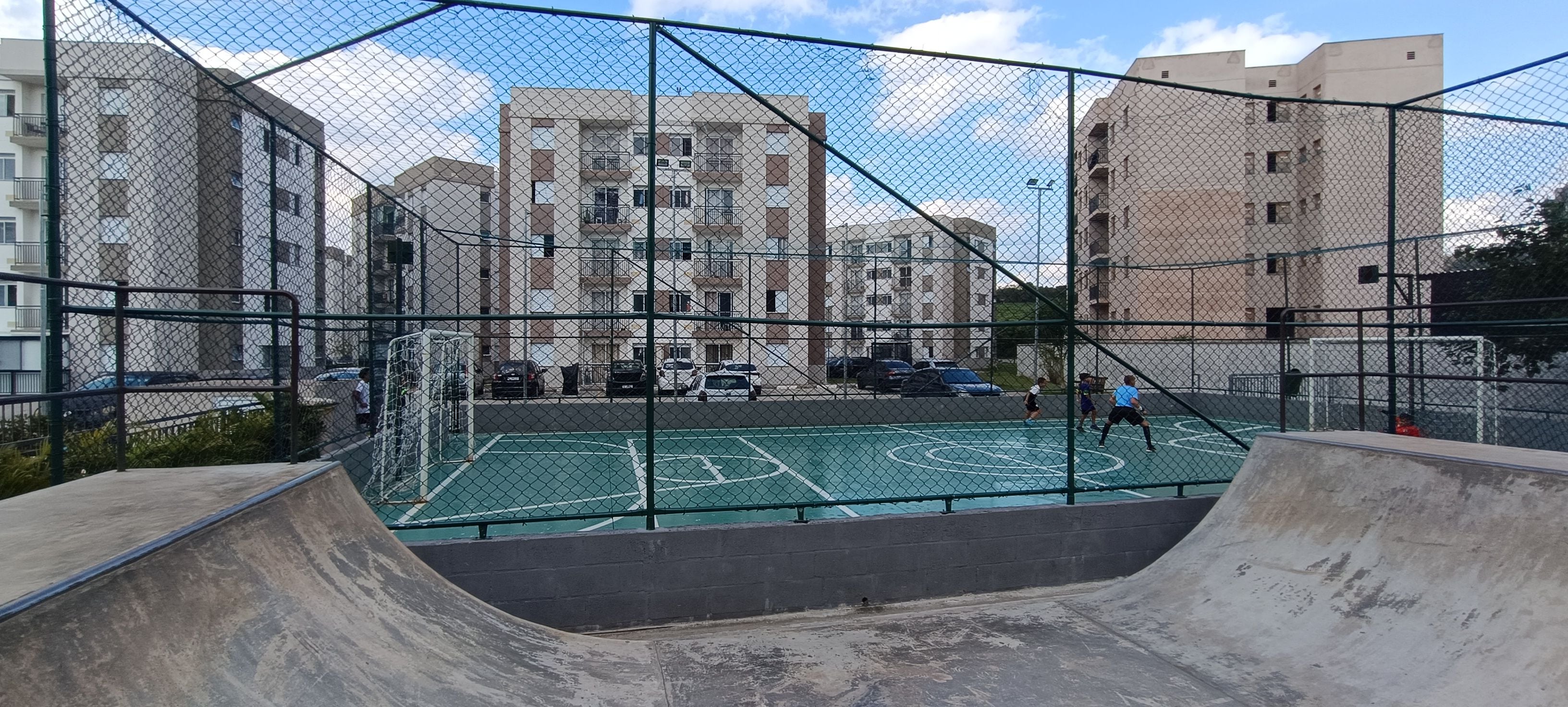 An outdoor basketball court surrounded by a chain-link fence, with residential buildings in the background.