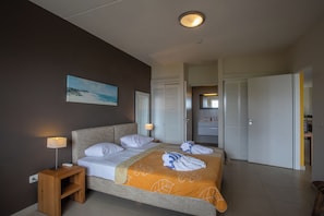 A hotel room with a bed, bedside lamps, a painting, a bathroom with a mirror, and a door. - Blue Bay Beachfront Apartment (Willemstad)