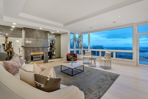 A spacious living room with a fireplace, a large window with a city view, and modern furniture. - Luxury 6BR West Van Villa and Sky-High Ocean Views (West Vancouver)