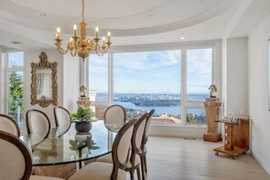 A dining area with a glass table, chairs, a chandelier, and a view of a cityscape through large windows. - Luxury 6BR West Van Villa and Sky-High Ocean Views (West Vancouver)