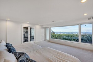 Ein Schlafzimmer mit einem großen Bett, einem Schrank mit Spiegel und Blick auf Grünflächen sowie auf die Stadt durch große Fenster.