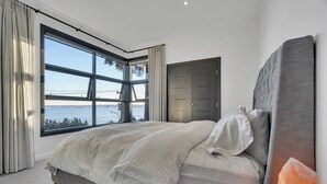 Quarto com uma janela ampla, cama com roupa de cama branca, cabeceira cinza e mesa de cabeceira de madeira.
