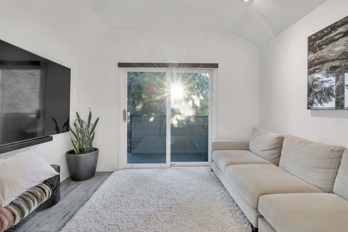 Un salon moderne avec un canapé, un téléviseur à écran plat, une plante en pot et une porte-fenêtre coulissante donnant sur un balcon.