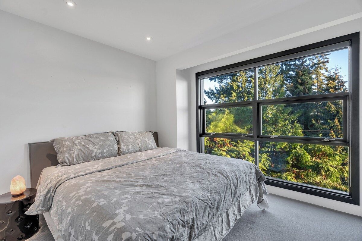 Quarto com uma janela grande, cama com roupa de cama cinzenta e mesa de cabeceira com luminária.