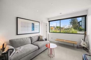 A modern living room with a grey sofa, a round table, a bench, and a large window offering a view of greenery. - Luxe 3BR Beachfront Home | Rooftop Terrace w/ BBQ (Vancouver)
