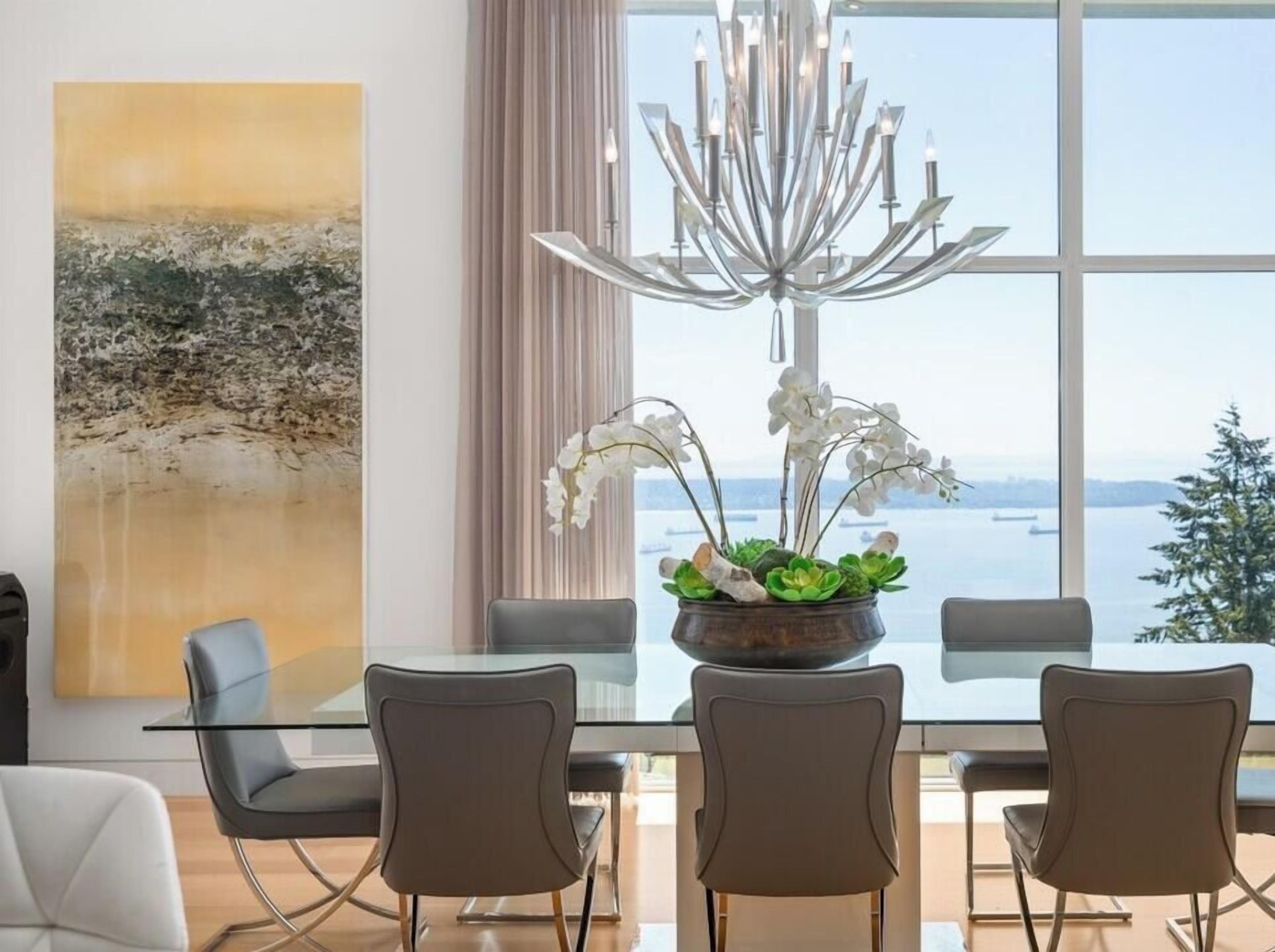 Un espace repas moderne avec une table en verre, quatre chaises, un lustre et une vue sur la mer.