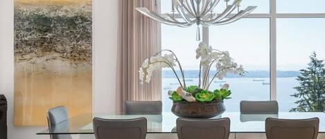 Un espace repas moderne avec une table en verre, quatre chaises, un lustre et une vue sur la mer.