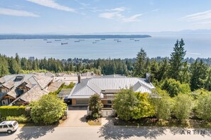 A residential area with a house featuring a sloped roof and a garage, surrounded by dense greenery and a view of the water with several boats. - Luxury 5BR Mansion | Pool, Sauna & Scenic Views (West Vancouver)