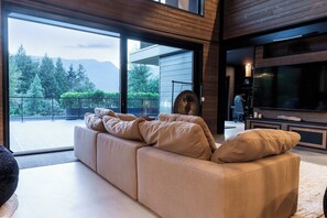 Ein modernes Wohnzimmer mit einer großen Couch, einem Flachbildfernseher und Blick auf eine gebirgige Landschaft durch große Fenster.