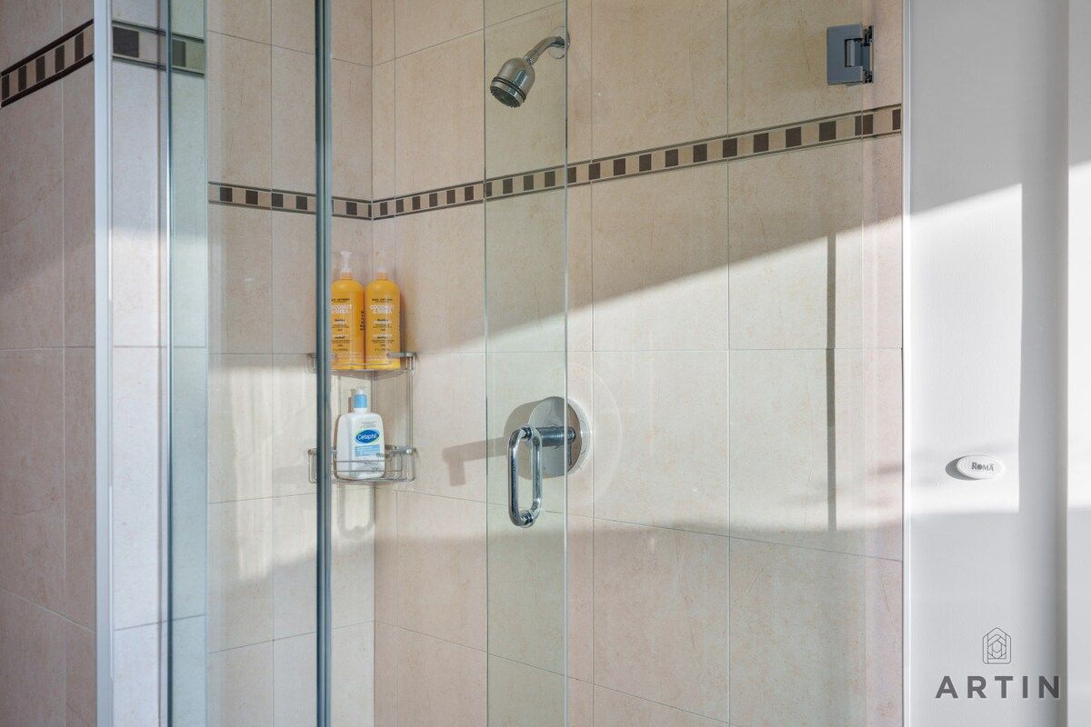 Ducha envuelta en vidrio, con cabezal de ducha de mano, repisa de vidrio con artículos de aseo personal y panel de control en la pared.