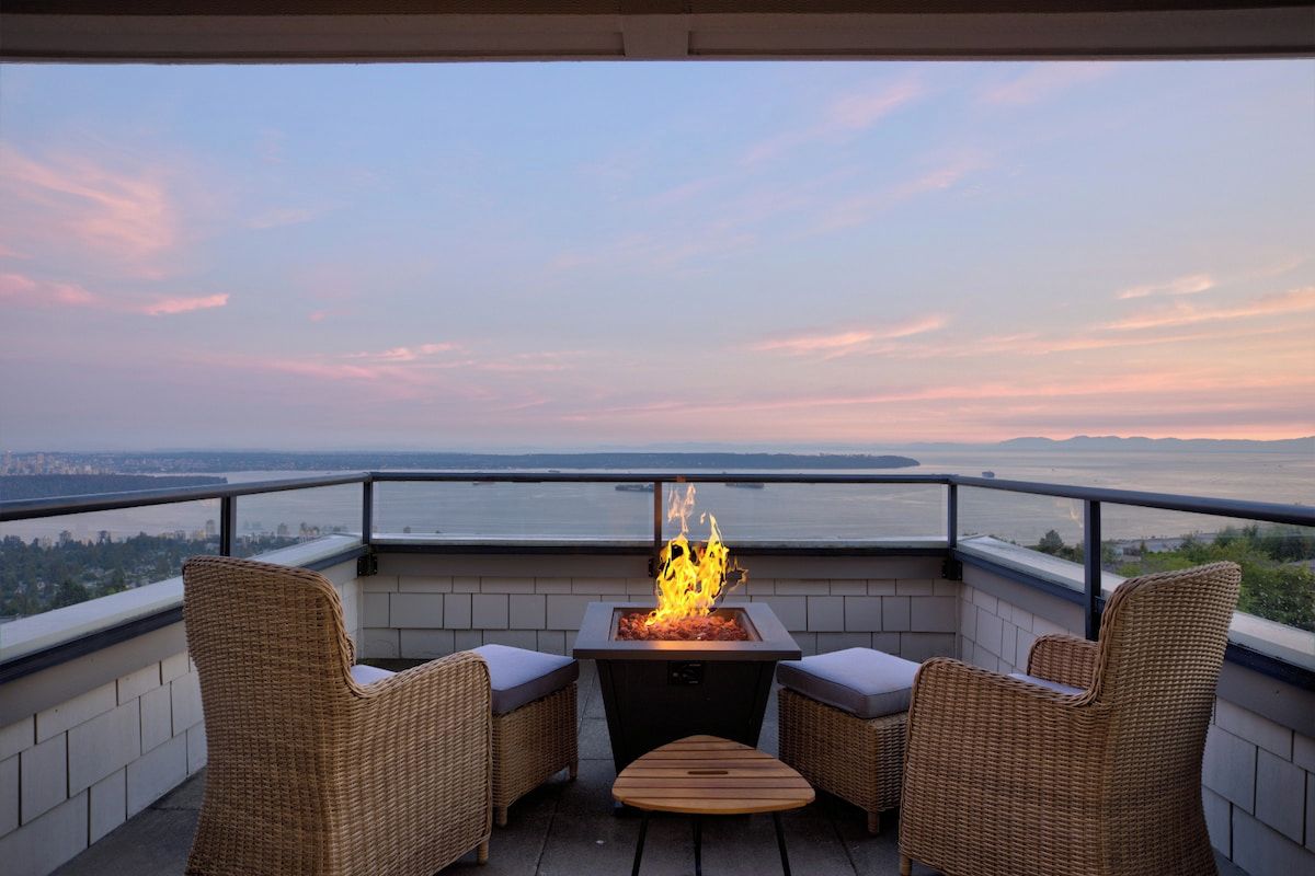 Ein Balkon mit Feuerstelle, zwei Korbstühle und Blick auf ein Gewässer sowie eine entfernte Skyline.