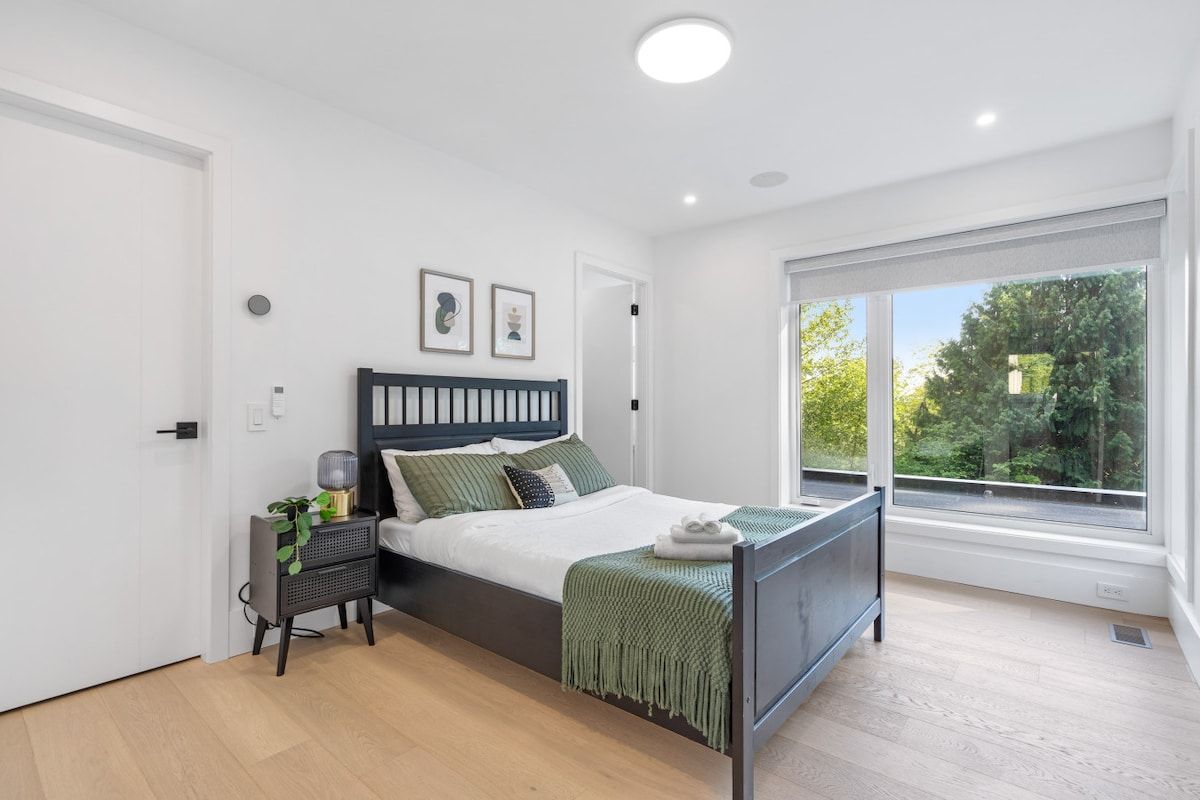 A modern bedroom with a large bed, a bedside table, a window with a view of greenery, and a ceiling light.