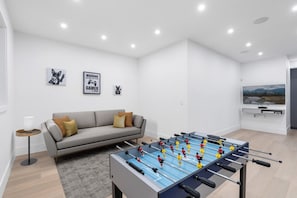 Ein modernes Wohnzimmer mit einem Kicker, einem grauen Sofa mit Kissen, einem kleinen Beistelltisch mit einer Lampe und einem flachbildfernseher an der Wand.