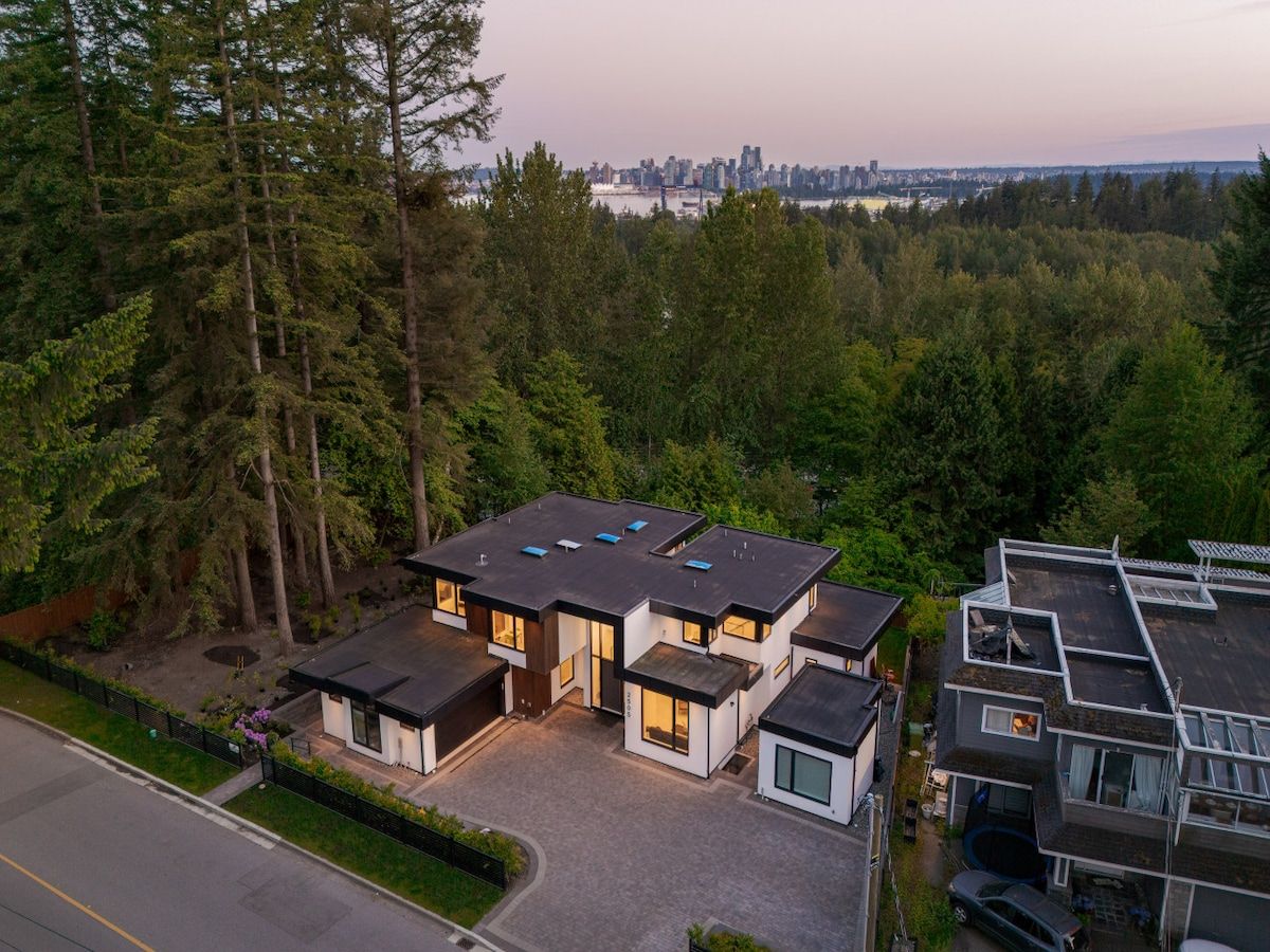 A modern two-story house with a flat roof, surrounded by dense forest.