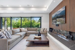 A modern living room with a sectional sofa, a coffee table, and a fireplace with a TV mounted on the wall. - Edgemont 5BR Designer Getaway with Home Theater (North Vancouver)
