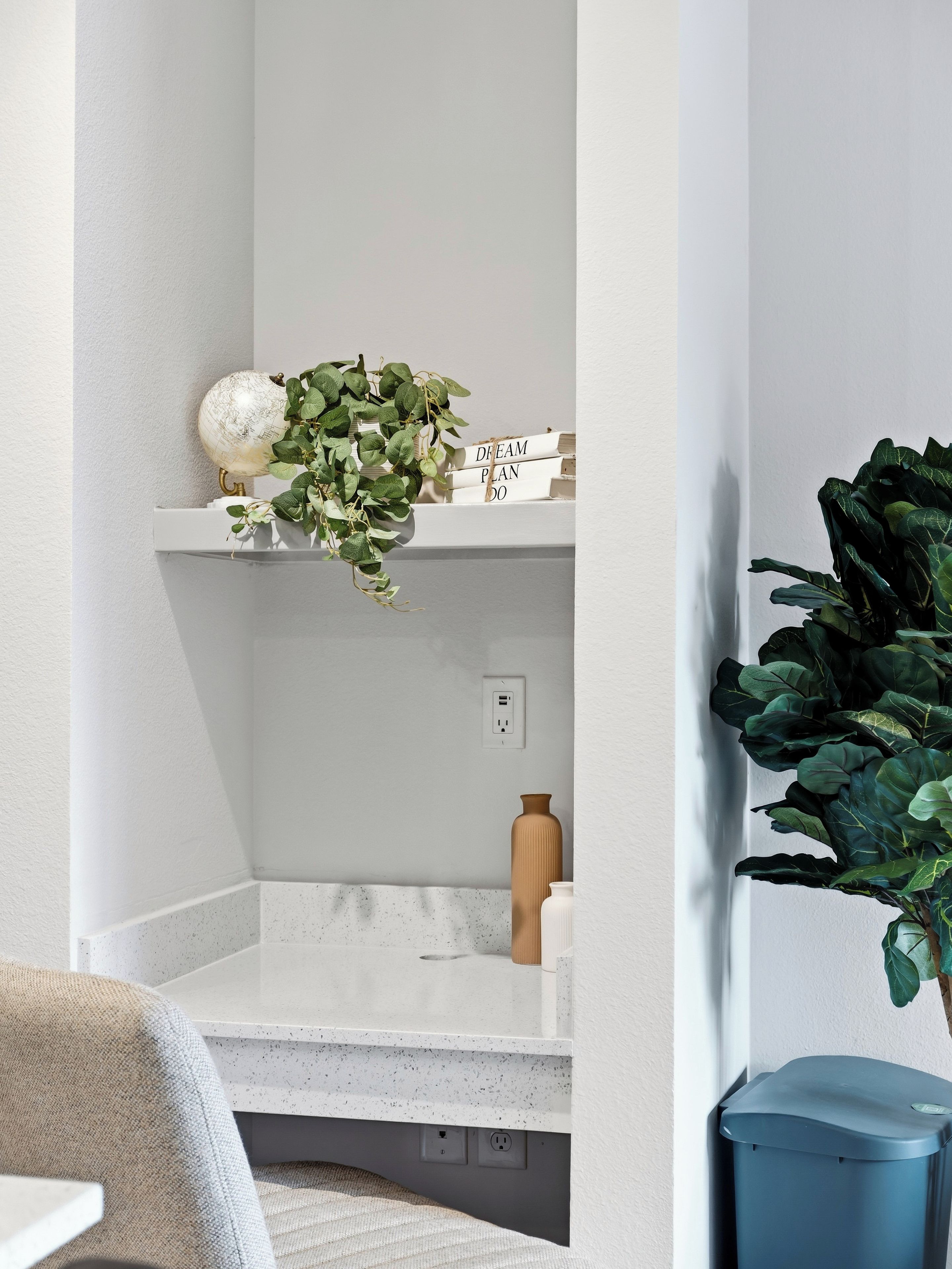 Una esquina de una habitación con una pared blanca, una estantería blanca con una planta, un pequeño jarrón y un cubo de basura azul.