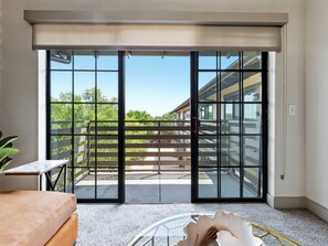 Una habitación con puerta de vidrio corrediza que da a un balcón exterior con vista a un edificio y árboles.