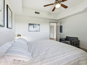 Un dormitorio con una cama grande, un ventilador de techo, un cuadro abstracto enmarcado y una puerta que da a otra habitación.