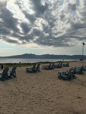 Une plage bordée de rangées de chaises et de tables Adirondack vertes, une rive sablonneuse et une vue sur l’eau et les montagnes lointaines, le tout sous un ciel nuageux.