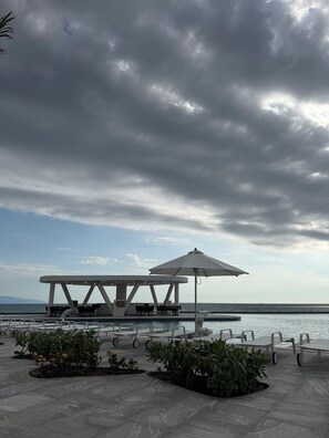 Uma área externa com mesas e cadeiras, um pavilhão coberto e vista para o mar.
