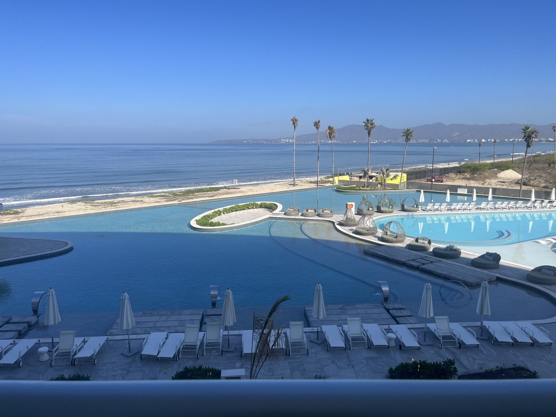 A resort pool with multiple sections, including a central area and a surrounding deck with lounge chairs and umbrellas.