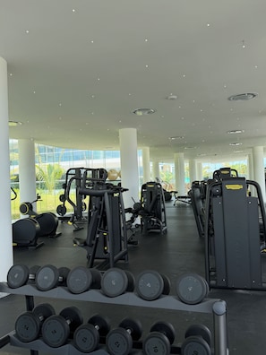 Un gimnasio con diverso equipo de ejercicio, incluyendo mancuernas y máquinas de pesas, y amplios ventanales que ofrecen luz natural.