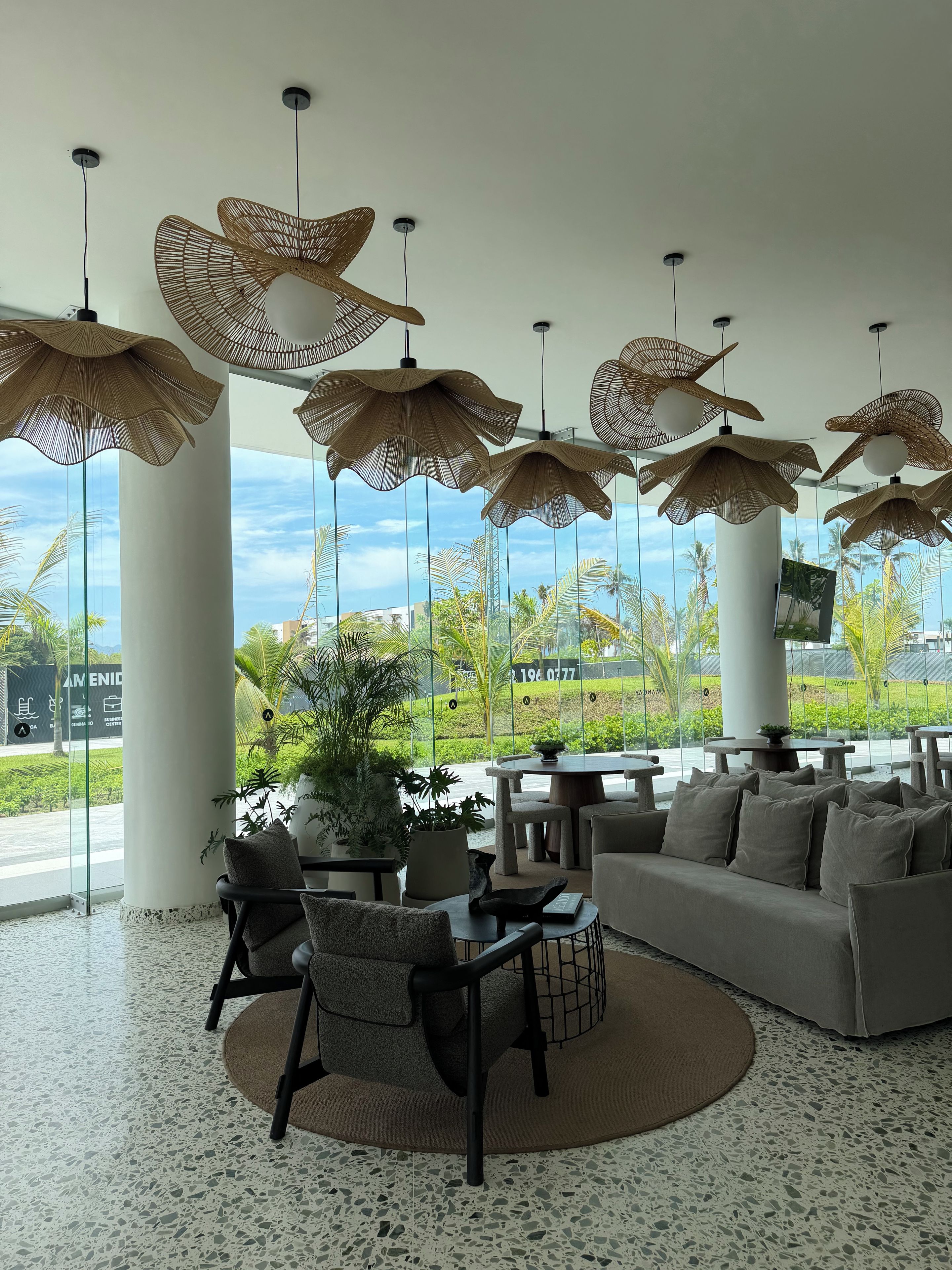 Un espace de détente moderne comprenant un canapé, des fauteuils et une table ronde, agrémenté de grandes fenêtres et de lampes suspendues en osier.