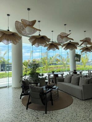 A modern lounge area with a sofa, chairs, and a round table, featuring large windows and hanging wicker lamps. - New 3BR beachfront apartment with incredible view and must see infinity pool. (Nuevo Nayarit)