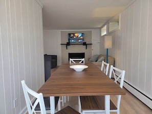 A dining area with a long wooden table, white chairs, a white bowl, and a television mounted on the wall. - Adorable 1-bedroom apartment in lovely Swampscott perfect for getaway (Swampscott)