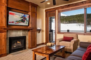 Una sala de estar acogedora con chimenea, un televisor montado en la pared y un amplio ventanal con vistas a un paisaje nevado.