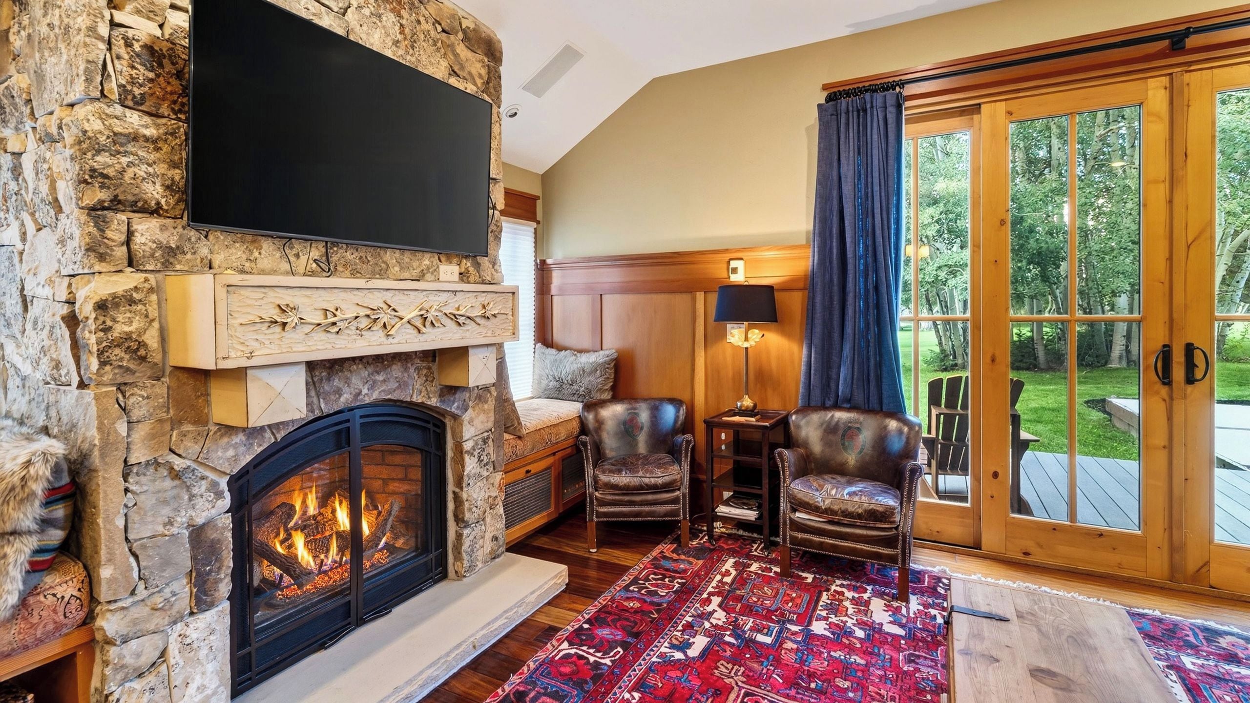 Une cheminée en pierre avec un téléviseur fixé au-dessus, des fauteuils en cuir, un tapis à motifs et une grande porte vitrée donnant sur l’extérieur.