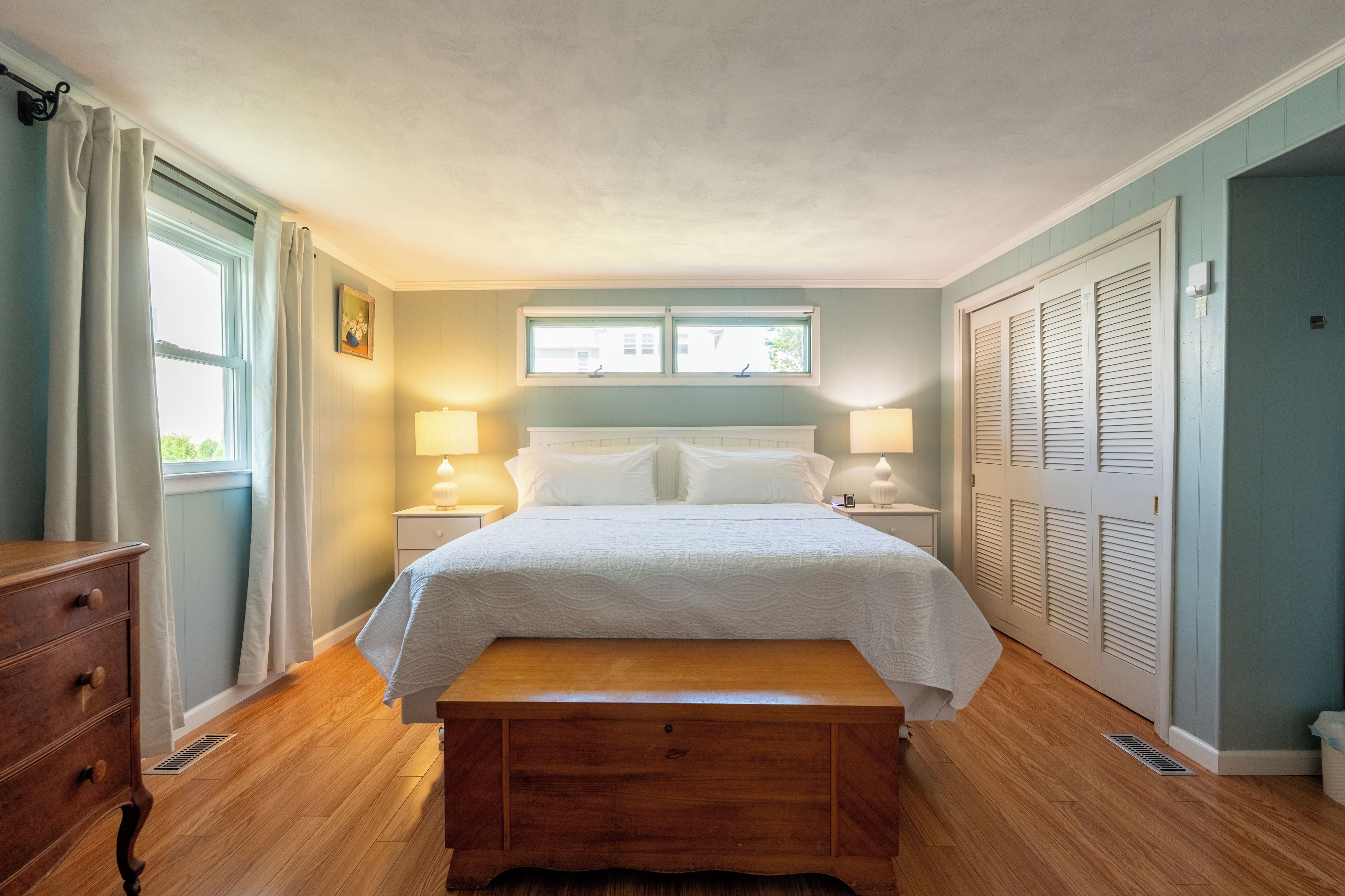 A bedroom with a bed, bedside lamps, a wooden bench, a window, and a door with louvered shutters.