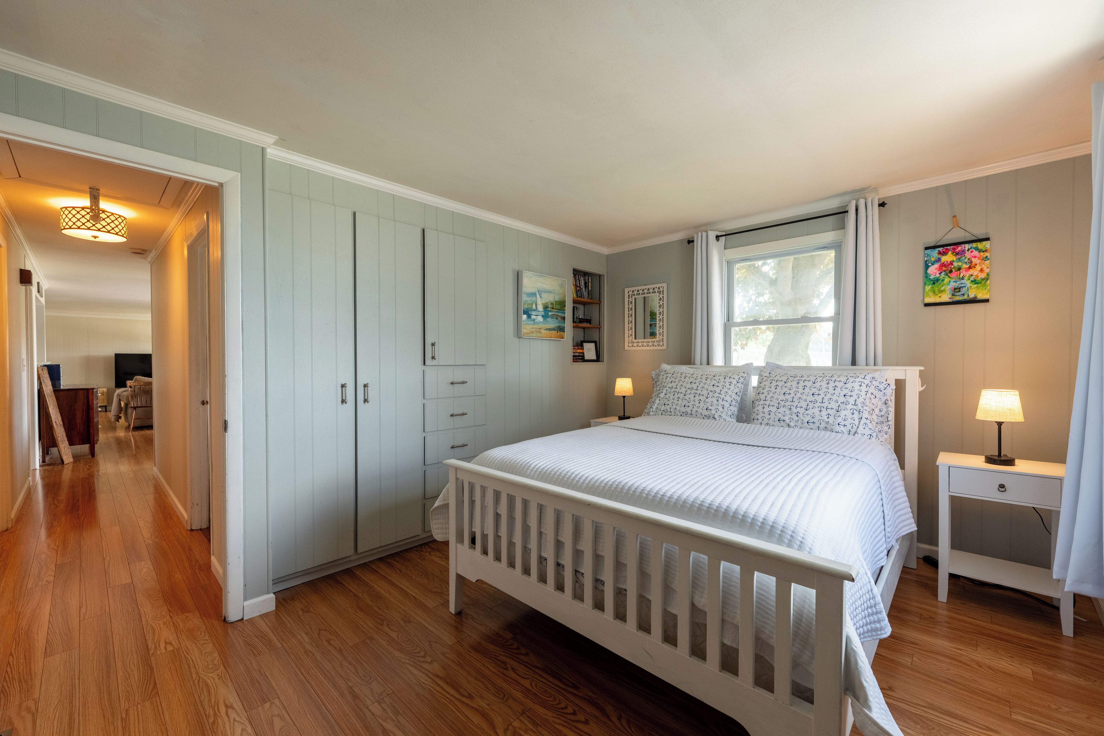 A bedroom with a white crib, a bedside table with a lamp, a window with curtains, and a door leading to another room.