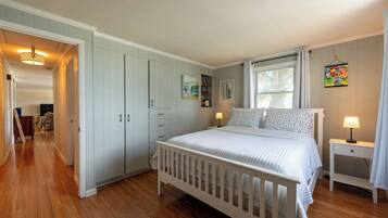 A bedroom with a white crib, a bedside table with a lamp, a window with curtains, and a door leading to another room.