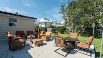 A covered outdoor patio with wicker furniture and a table, surrounded by a wooden deck and a house with a solar panel roof.