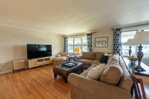 A living room with a flat-screen TV, a sofa, a coffee table, and a patterned curtain. - Beach House w/ Spacious Yard, Walk to Beach with AC. 10 Minutes to Mystic! (Groton)